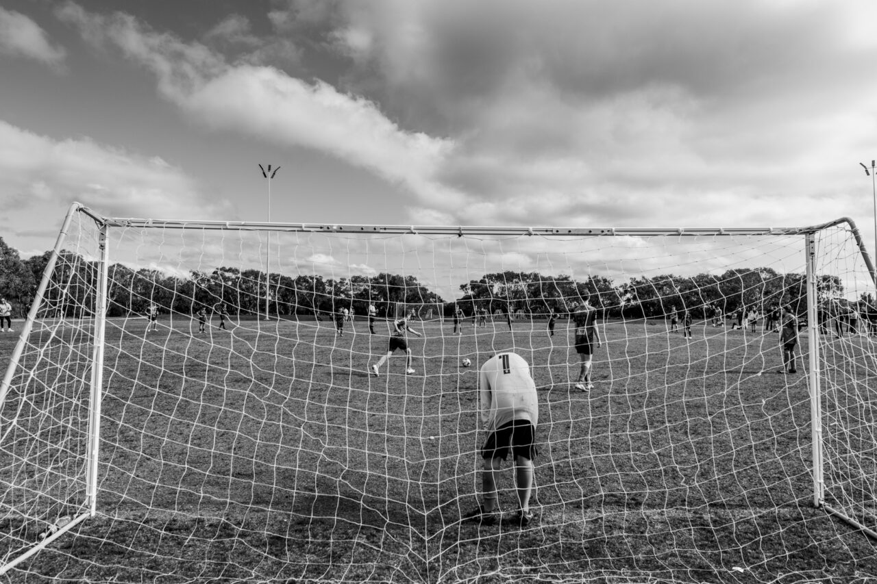 View through a soccer goal frame across the field under a dramatic sky, black and white