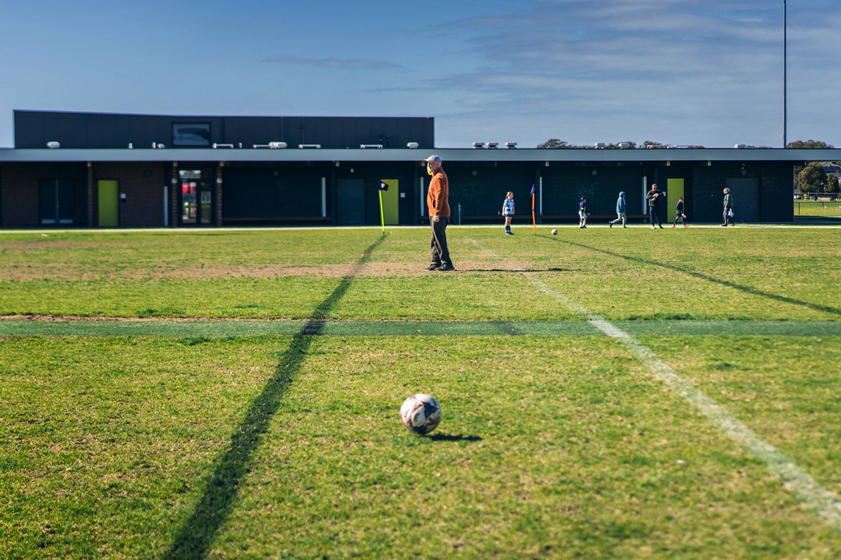 Official standing on a soccer field with a ball in the foreground, community centre behind