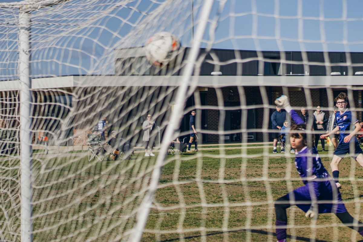 Soccer ball hitting the back of the net, shot through the goal net with the goal scorer and the keeper visable