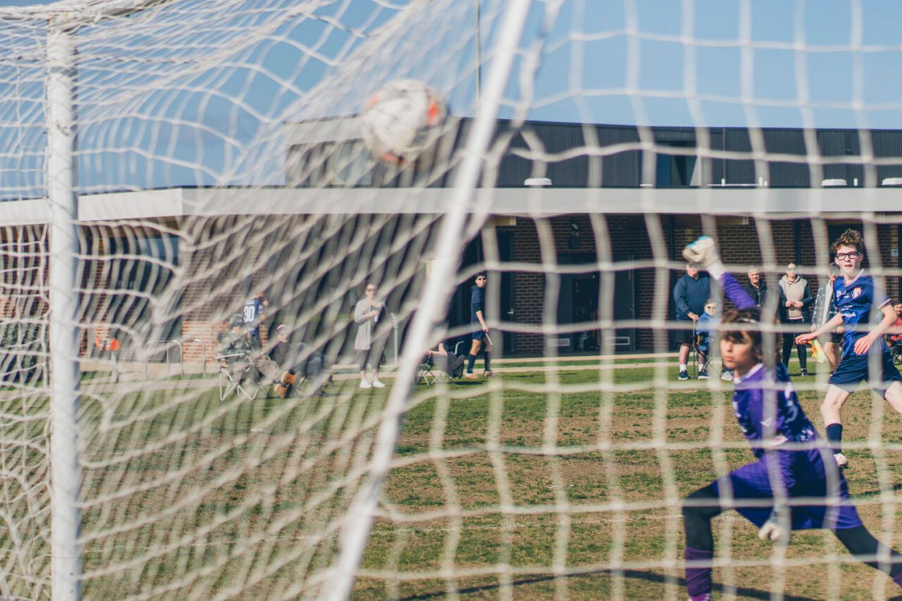 A soccer ball flying into the goal net at an AUFC under 15s match with sideline supporters watching