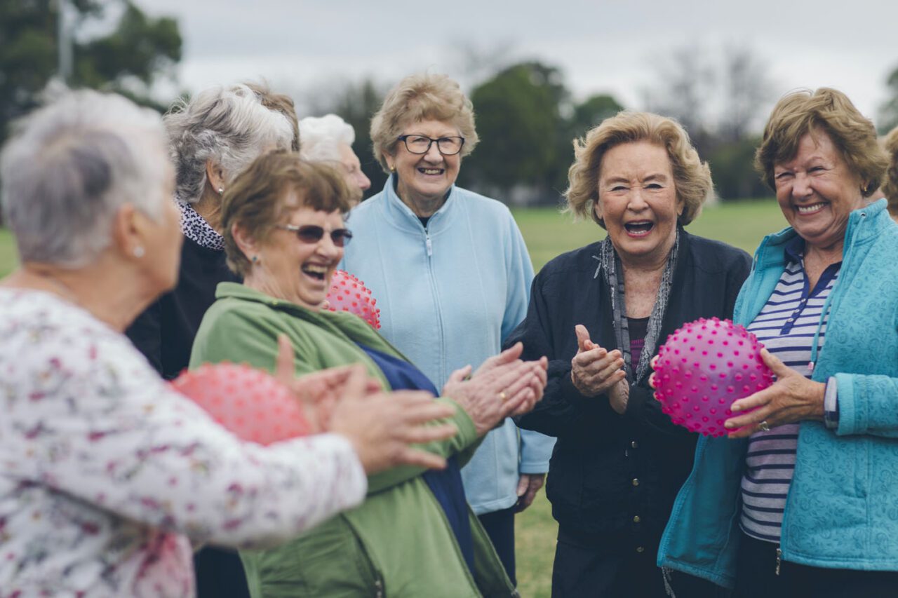 A group of senior women laughing together while holding textured exercise balls outdoors