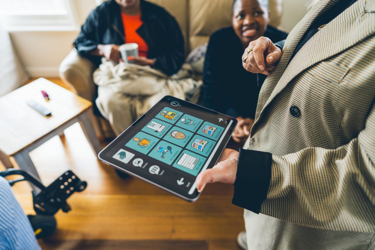 Support worker holding a tablet showing a communication app while two participants look on from the couch