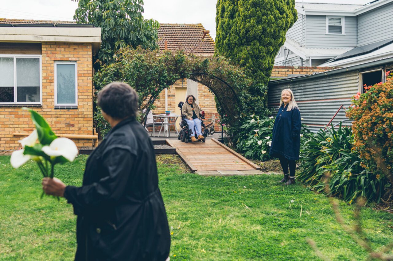 Woman in a wheelchair coming down a garden ramp through a rose arch while a support worker waits and another carries calla lilies