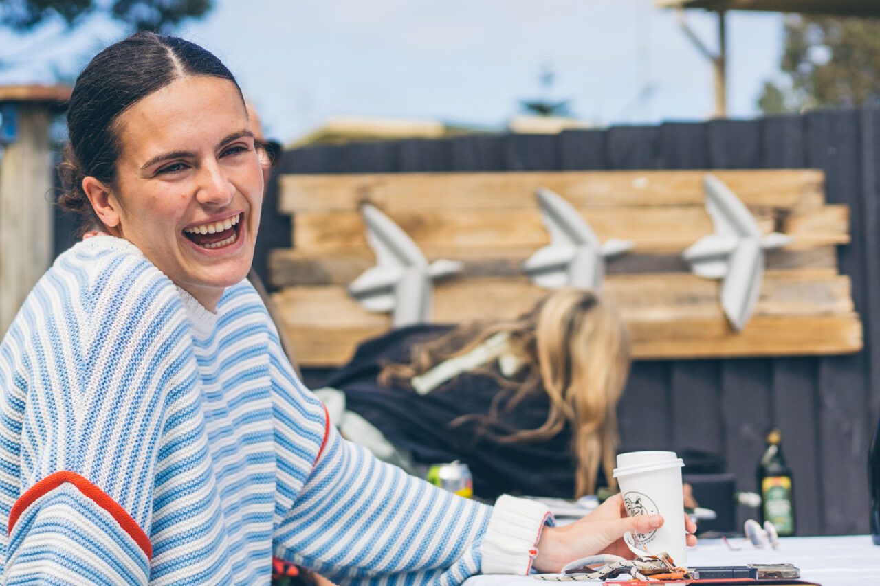 Woman in a blue striped jumper laughing with a takeaway coffee at an outdoor table